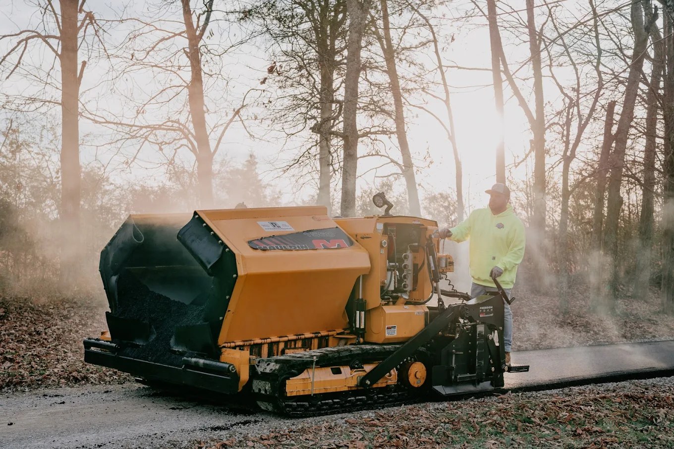American Asphalt paver and operator laying fresh hot-mix asphalt at golden hour